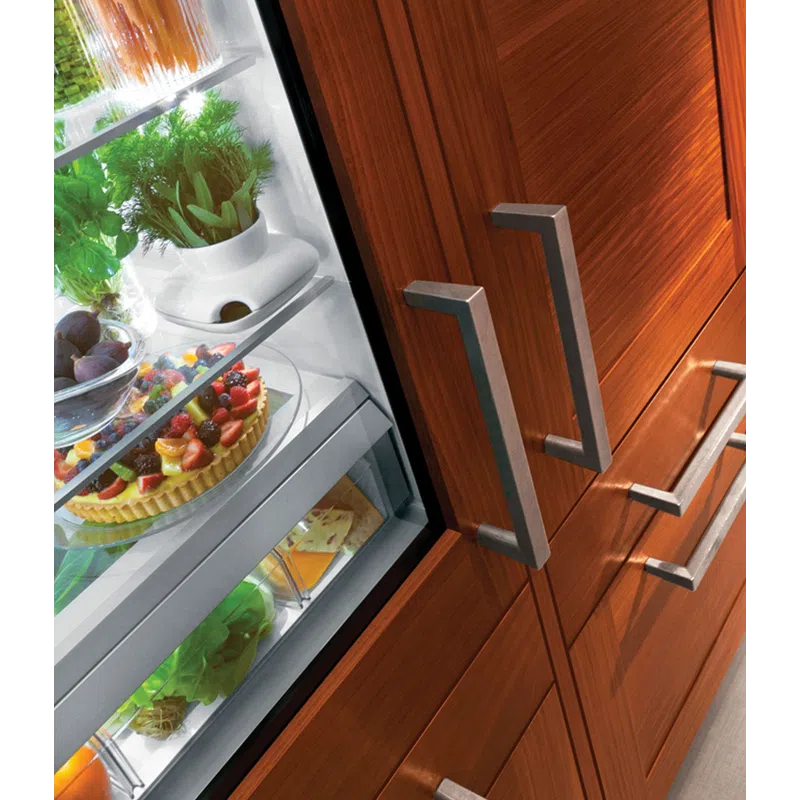 Open refrigerator door revealing fresh herbs, fruits, and a fruit tart inside wooden kitchen cabinets.