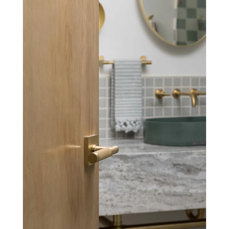 Partially open wood door with brass handle showing modern bathroom with round mirror and green sink.