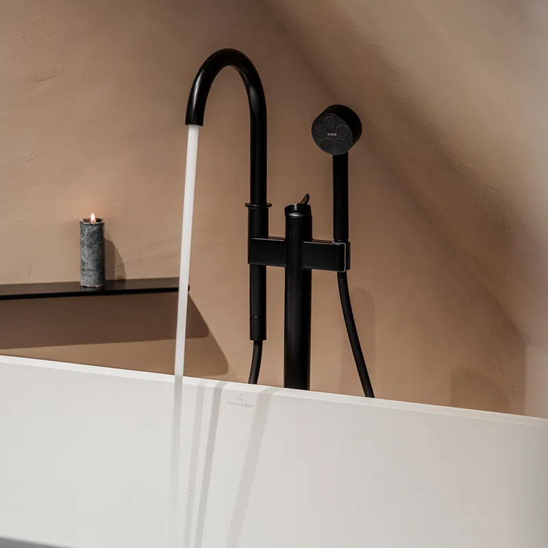 Modern black freestanding bathtub faucet running water with a lit candle on a beige wall shelf.