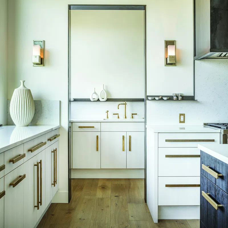 Modern kitchen with white cabinetry, gold handles, wooden floor, and decorative vases by the sink area.