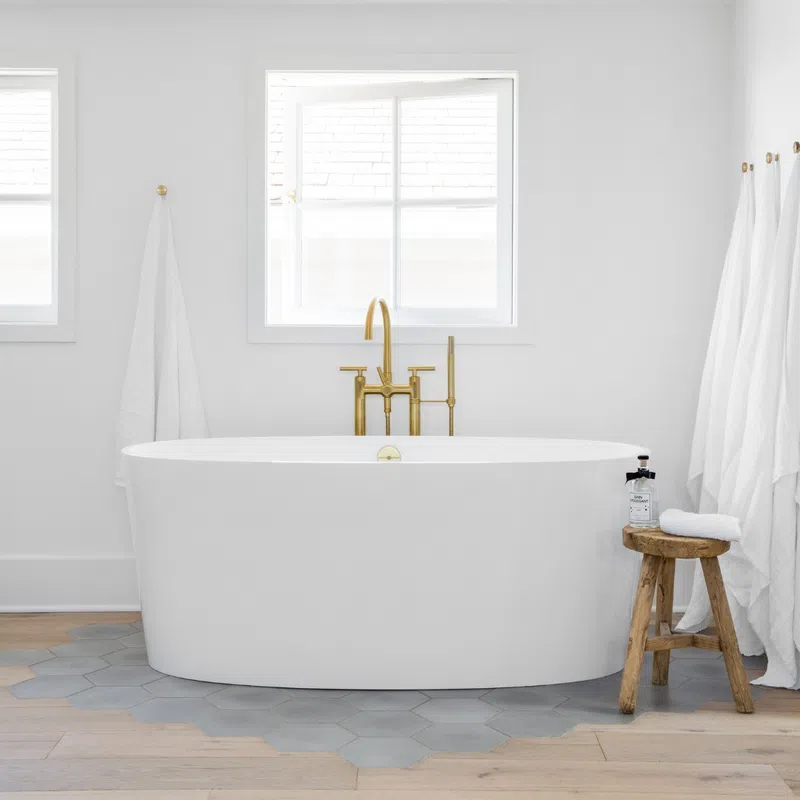 Modern white freestanding bathtub with gold faucet under a window, white towels, and wooden stool on hex tile floor.