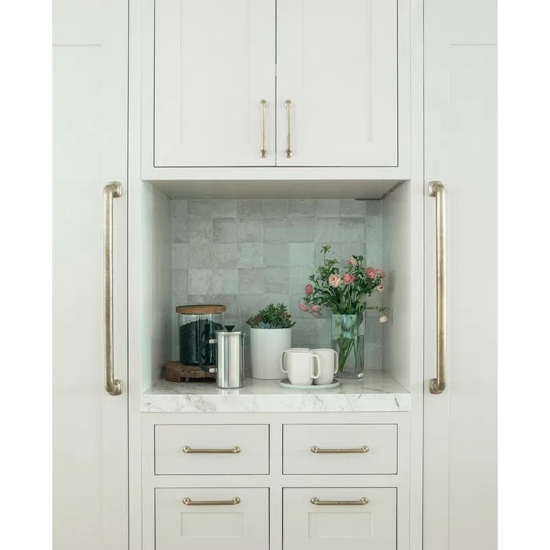 Minimalist kitchen nook with marble countertop, white cabinets, coffee accessories, plants, and a vase of flowers.