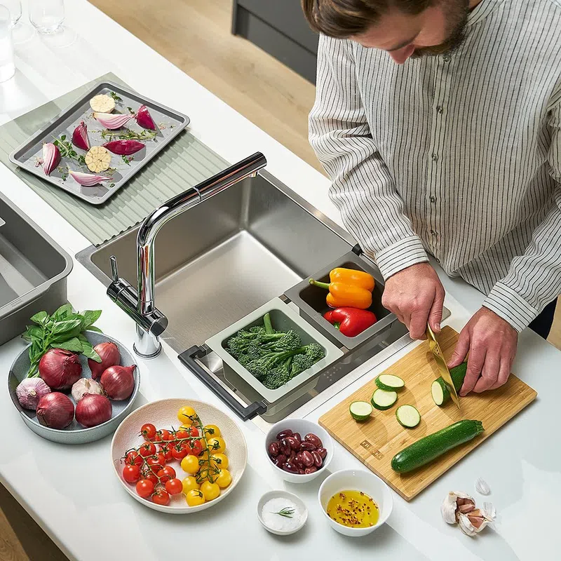 Person slicing zucchini on wooden board with various fresh vegetables and kitchen sink nearby in modern kitchen