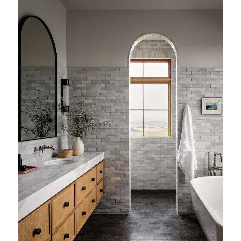 Modern bathroom with marble countertop, wood cabinetry, arched window, freestanding tub, and gray brick walls.