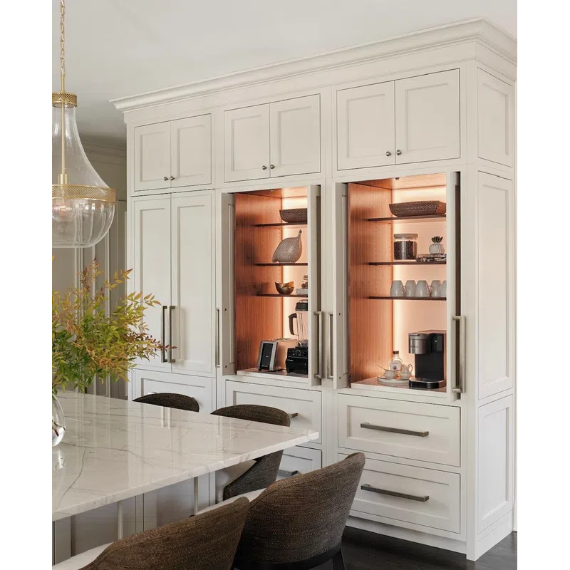 White kitchen cabinets with built-in warm-lit shelves displaying decor and coffee appliances beside a marble countertop.