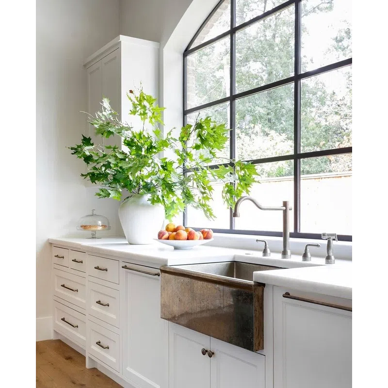 Bright white kitchen with large arched window, rustic farmhouse sink, green leafy plant, and bowl of fruit on the counter.
