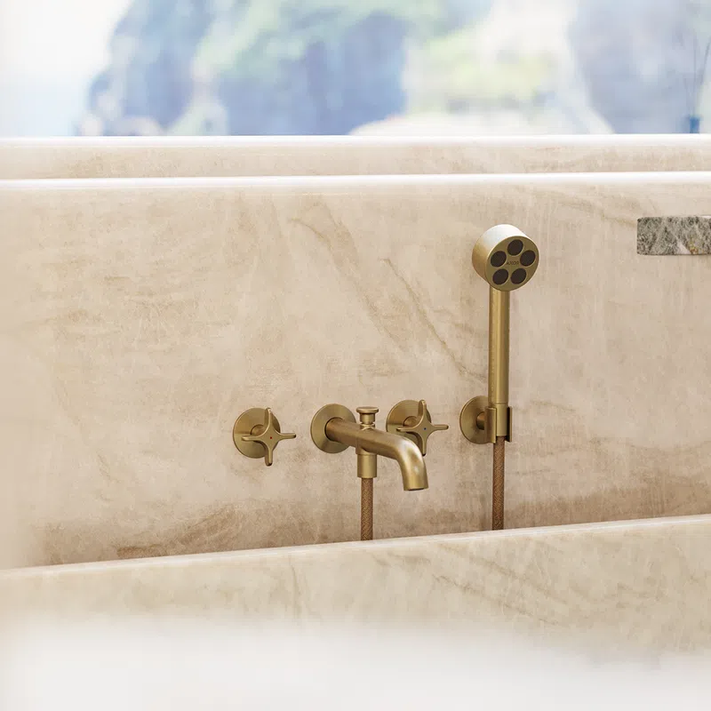 Modern brass bathtub faucet and handheld shower mounted on beige marble surface with soft natural background.