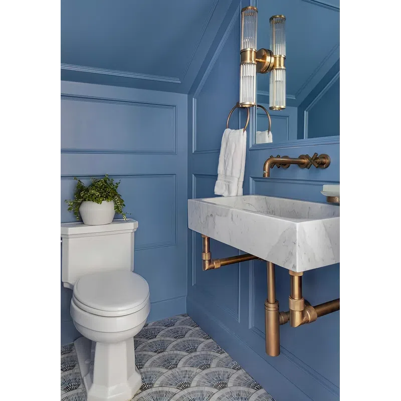 Modern blue bathroom with white toilet, marble sink, brass fixtures, patterned floor tiles, and a potted plant.