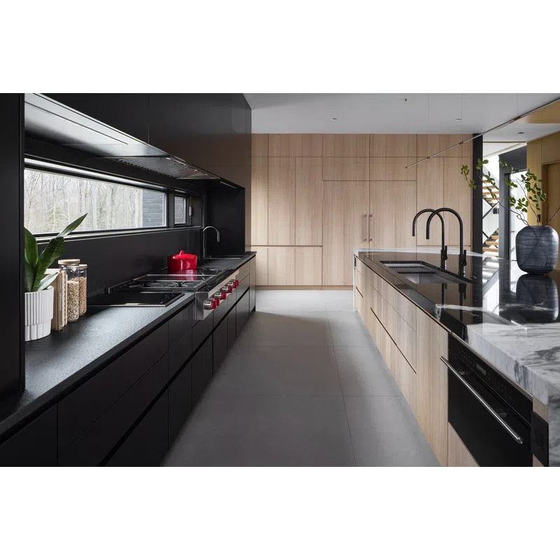 Modern kitchen with black and light wood cabinetry, black countertops, marble island, and large window overlooking trees.