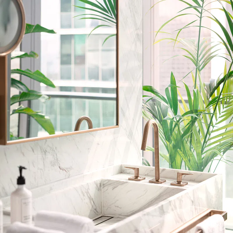 Modern bathroom sink with marble countertop, gold faucet, large mirror, and green plants by window.