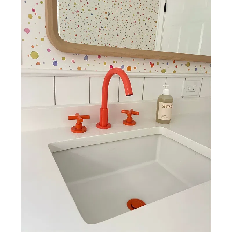 Modern white bathroom sink with vibrant orange faucet and handles, colorful dotted wallpaper, and liquid soap dispenser.