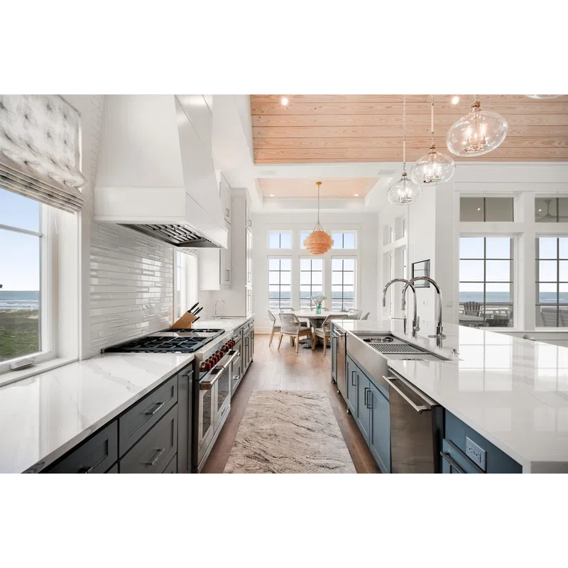 Bright modern kitchen with marble countertops, blue cabinetry, wooden ceiling, and ocean views through large windows.
