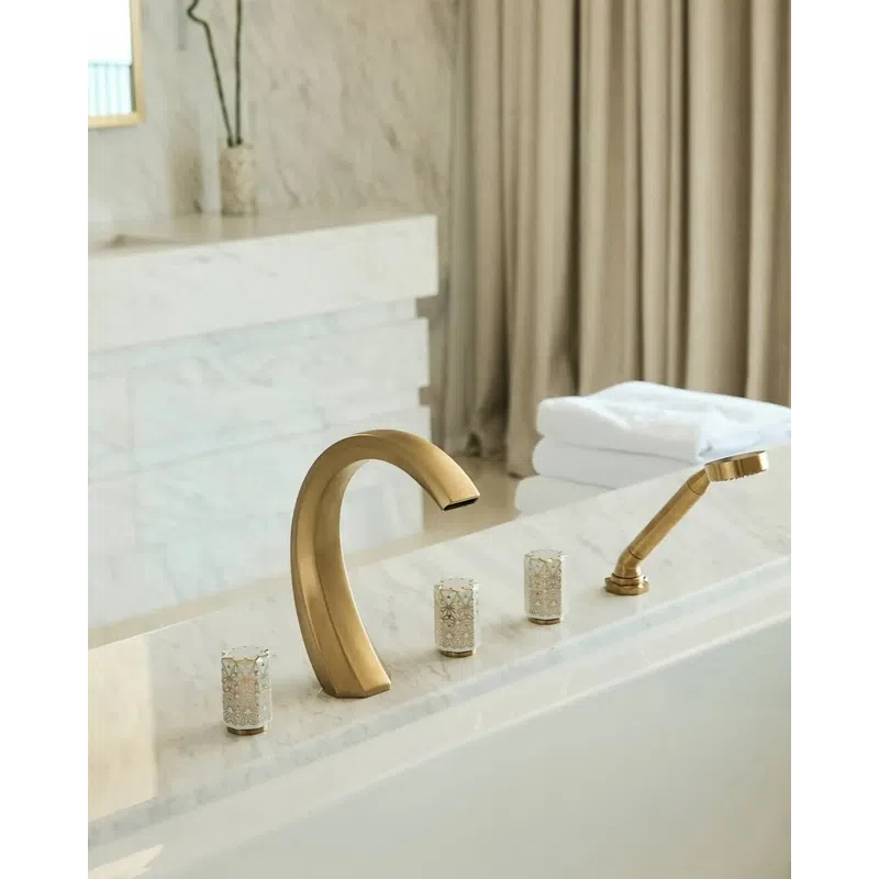 Modern gold bathroom faucet and handheld shower on white marble bathtub with folded towels in background.