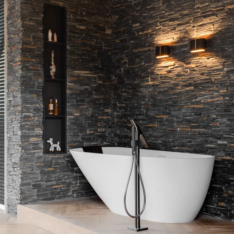 Modern bathroom with freestanding white tub, black stone wall, built-in shelves, and warm wall sconces lighting