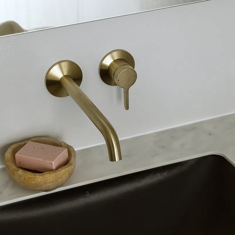 Modern brass wall-mounted faucet over marble countertop with brown sink and stone soap dish with pink soap.