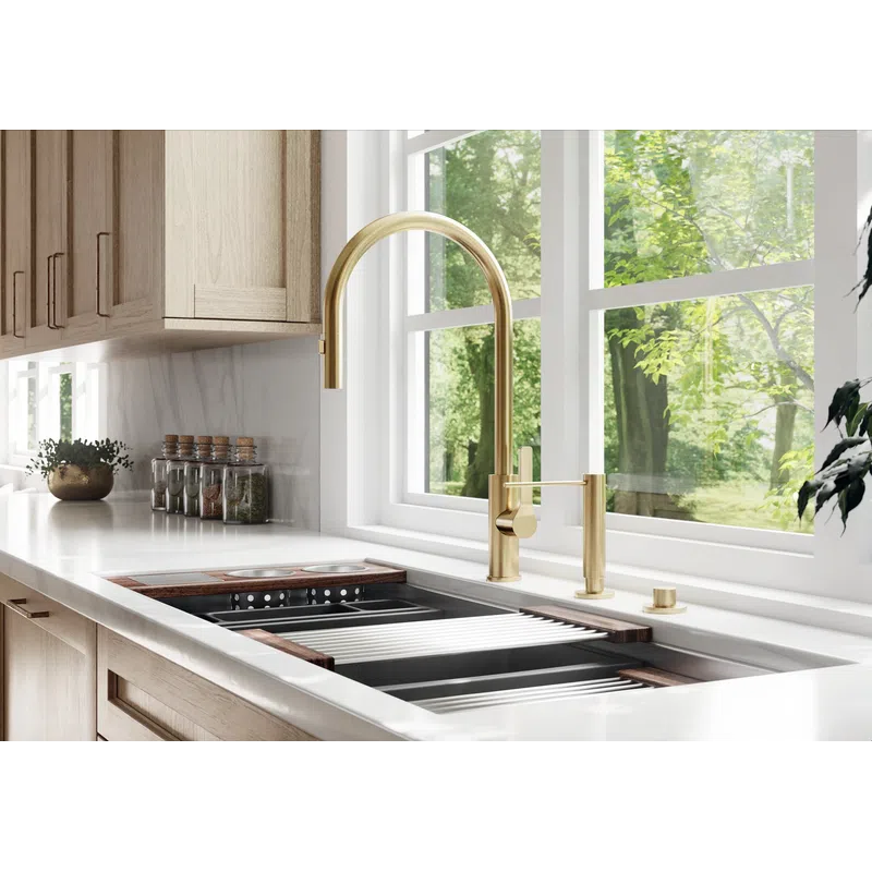 Modern kitchen sink with gold faucet, wooden cabinets, white countertop, and large window overlooking greenery.