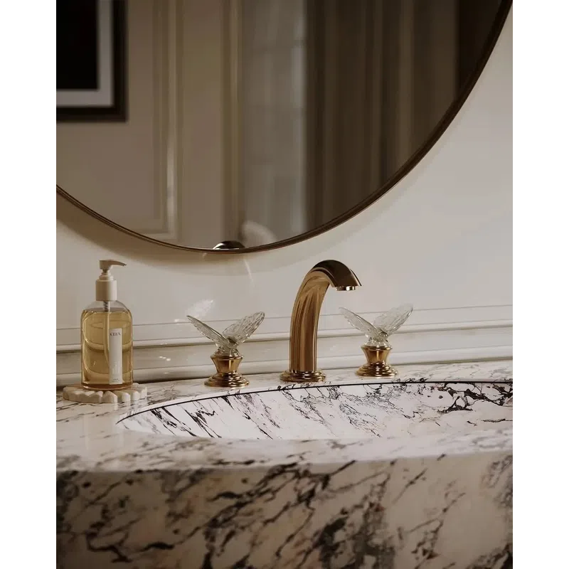 Marble bathroom sink with gold faucet and crystal butterfly handles, round mirror and liquid soap dispenser.