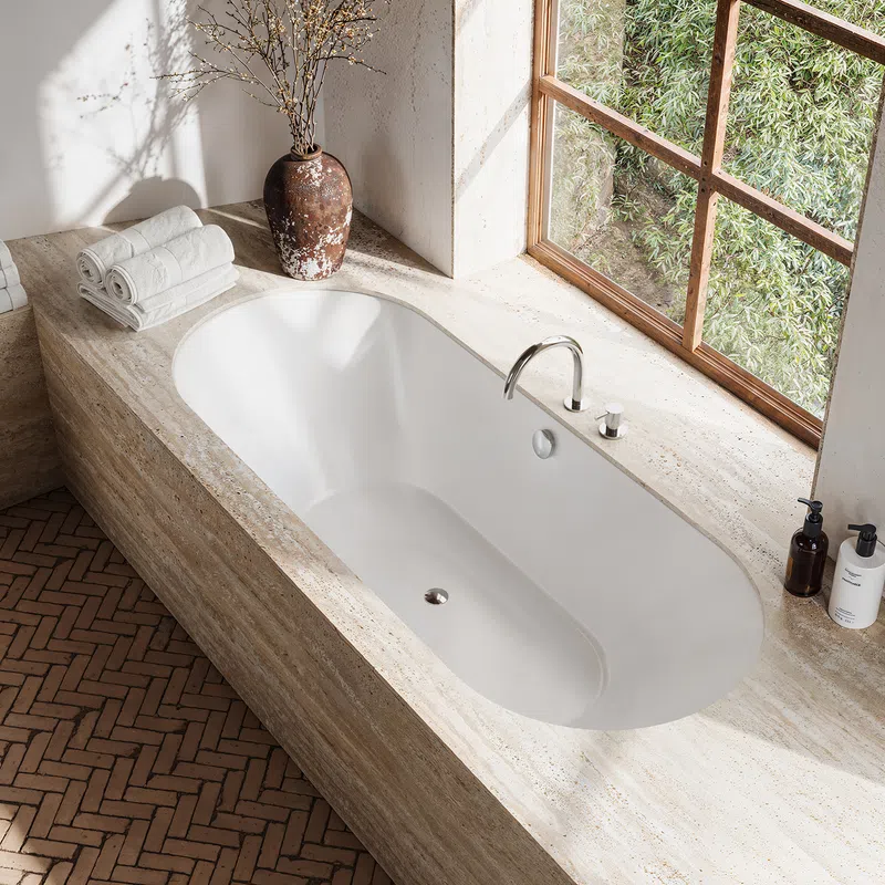 Modern bathroom with white built-in bathtub, natural stone surround, window, plants, and bath accessories.