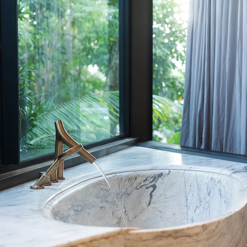 Modern marble bathroom sink with running water faucet next to large window overlooking greenery.