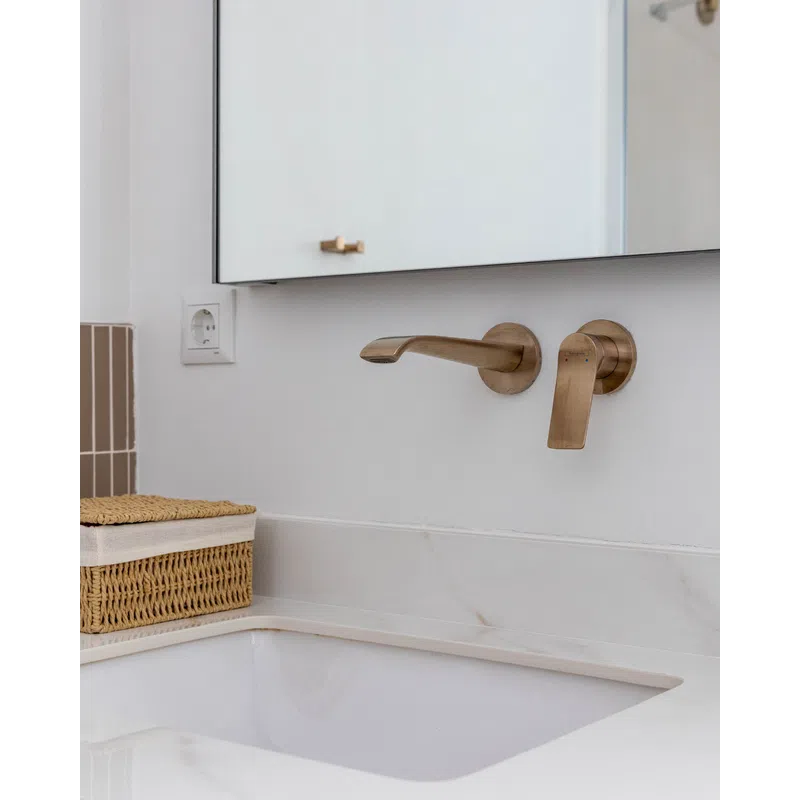 Modern bathroom sink with wall-mounted brushed brass faucet and woven storage basket on marble countertop.