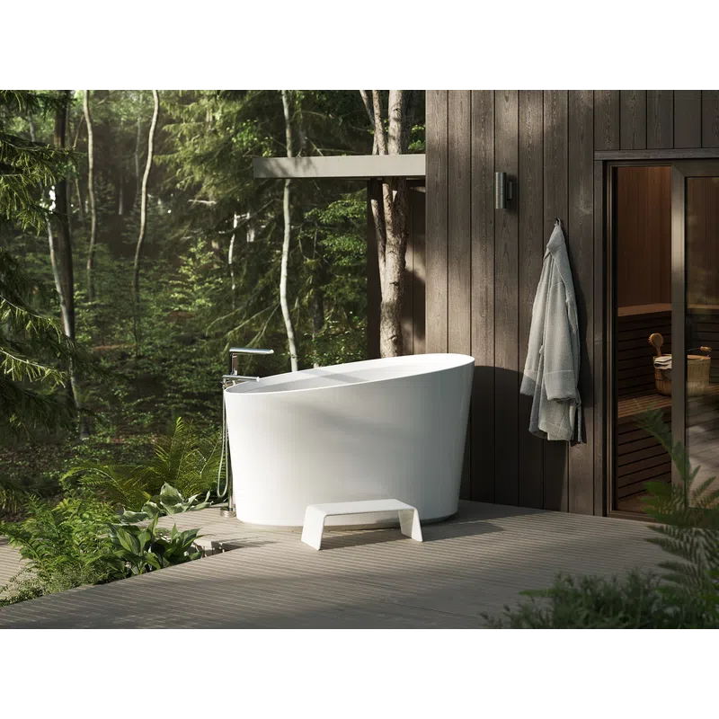 Modern white freestanding bathtub on wooden deck surrounded by forest greenery and attached sauna room.