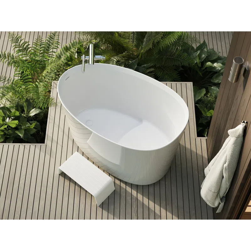 Modern white freestanding bathtub on wooden deck surrounded by green plants with towel hanging nearby