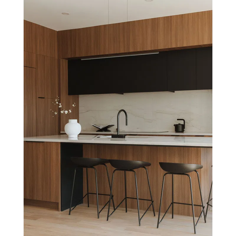 Modern kitchen with wood cabinets, marble countertop, black stools, and minimalist decor with white vase and flowers.