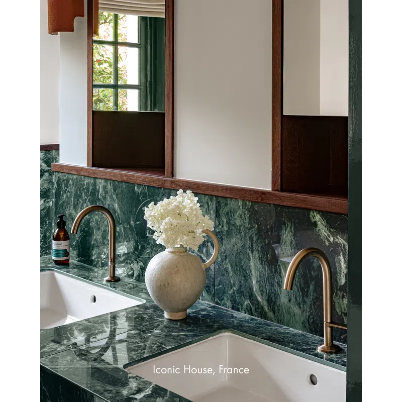 Modern bathroom with green marble countertop, dual brass faucets, white sinks, and a decorative white flower vase.