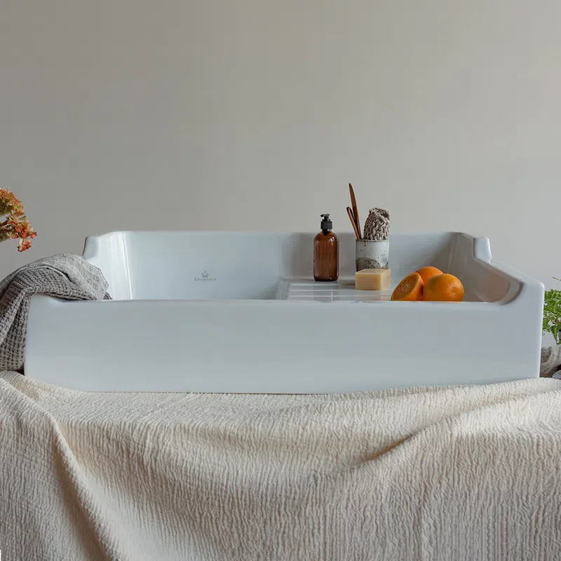 White ceramic bathtub tray with soap, brown bottle, brush, and oranges on a beige textured cloth.