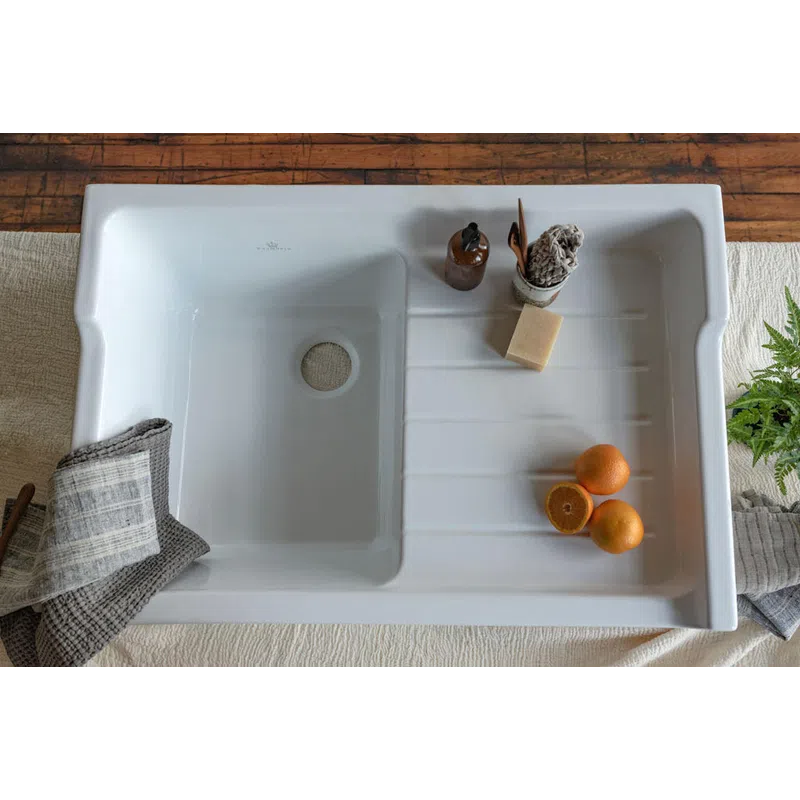 White ceramic kitchen sink with oranges, soap, and cloths on a wooden and fabric surface.