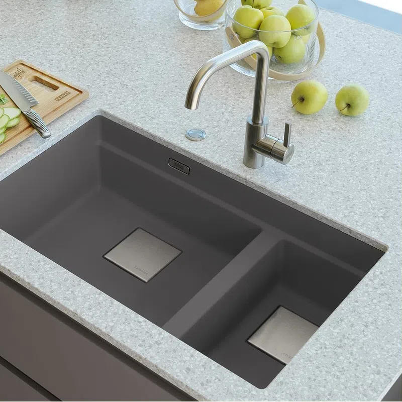 Modern double-basin gray kitchen sink with stainless steel faucet on light granite countertop with green apples and cutting board.