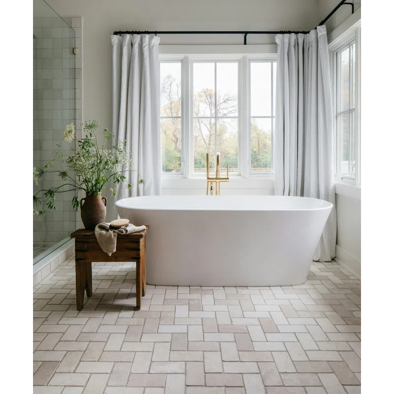Bright bathroom with freestanding white bathtub, wooden stool, vase of flowers, and large window with white curtains.