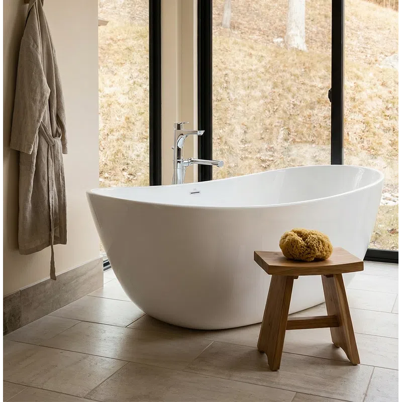 Modern white freestanding bathtub next to large windows with natural scenic view and wooden stool with bath sponge.
