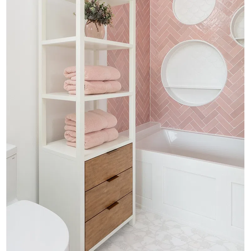 Modern bathroom with white shelving unit holding pink towels, wood drawers, pink tiled wall, and white bathtub.
