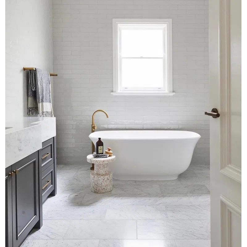 Modern bathroom with freestanding white tub, brass faucet, gray cabinets, marble floor, and a window above bathtub.