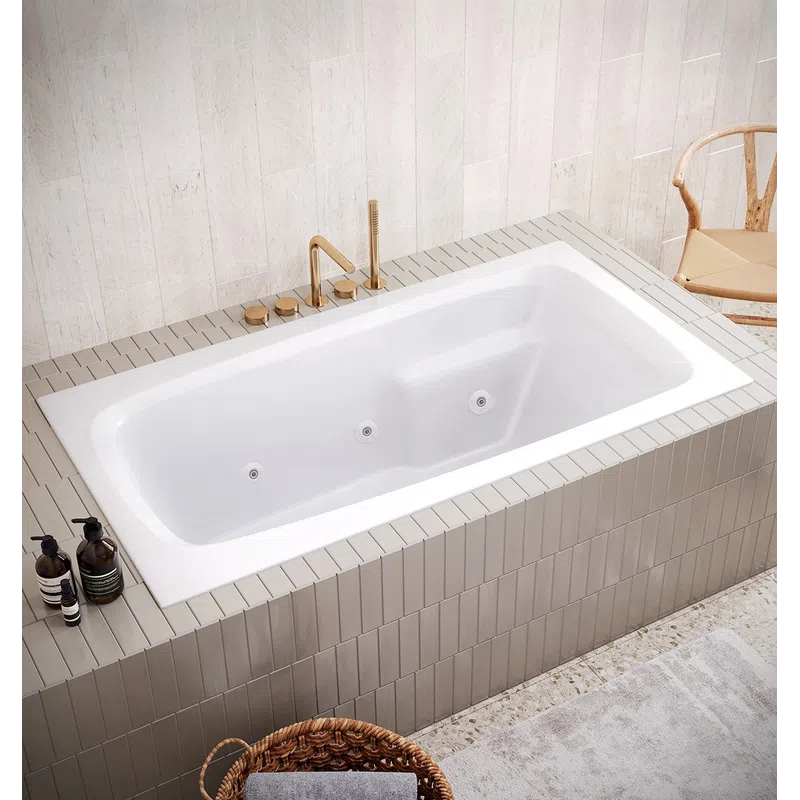 Modern white built-in bathtub with golden fixtures in a beige tiled bathroom with wicker basket and wooden chair.
