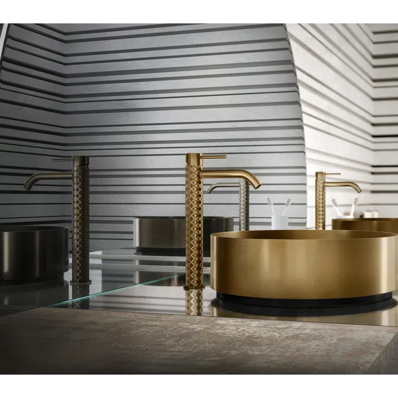Modern bathroom sink with a gold faucet and round gold basin against a gray striped wall and mirror.