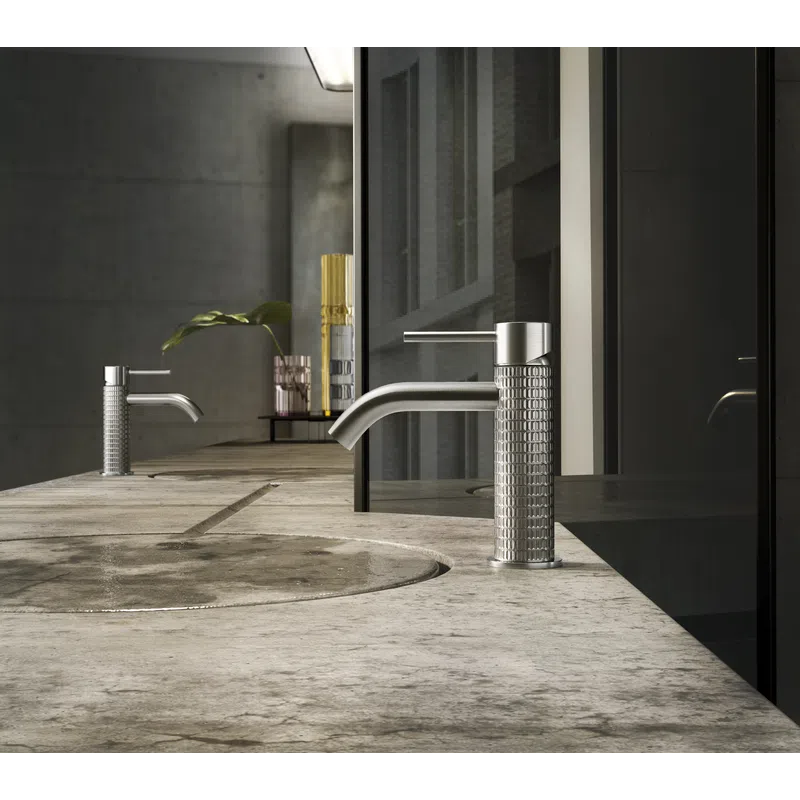 Modern bathroom sink with textured stainless steel faucet on a stone countertop with reflective dark glass backsplash.