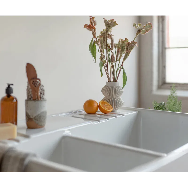 A white bathtub with dried flowers in a vase and oranges on the edge, next to soap and a brown bottle.