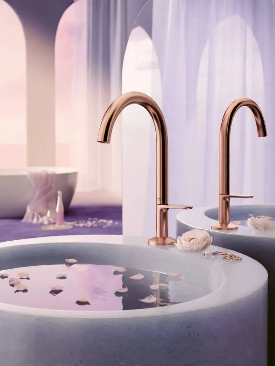 AXOR rose gold widespread faucets over pale stone vessel sinks beneath sheer purple curtains.