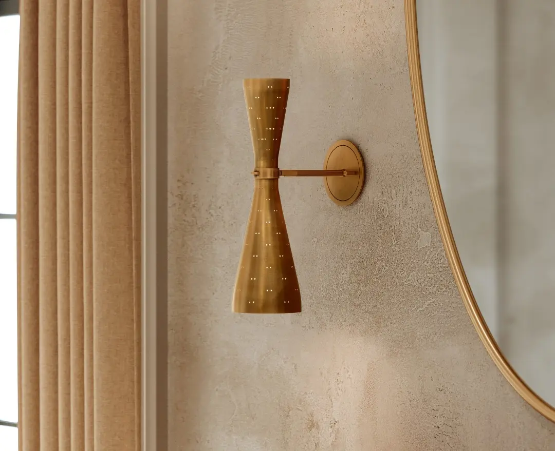 Modern brass wall sconce with perforated design mounted on textured beige wall beside large curved mirror.