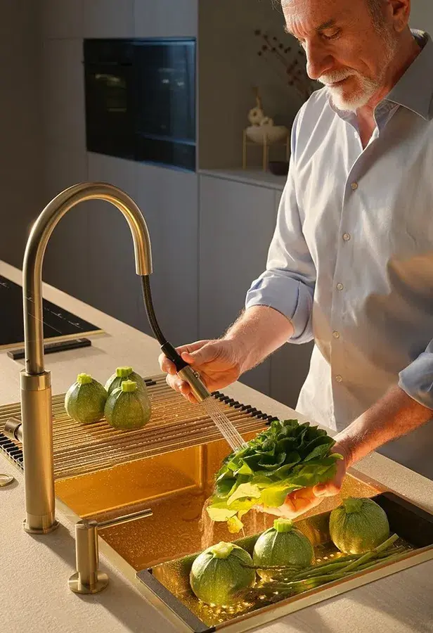 Man rinsing fresh vegetables under a brushed gold Franke pull-down kitchen faucet over a stainless steel sink.
