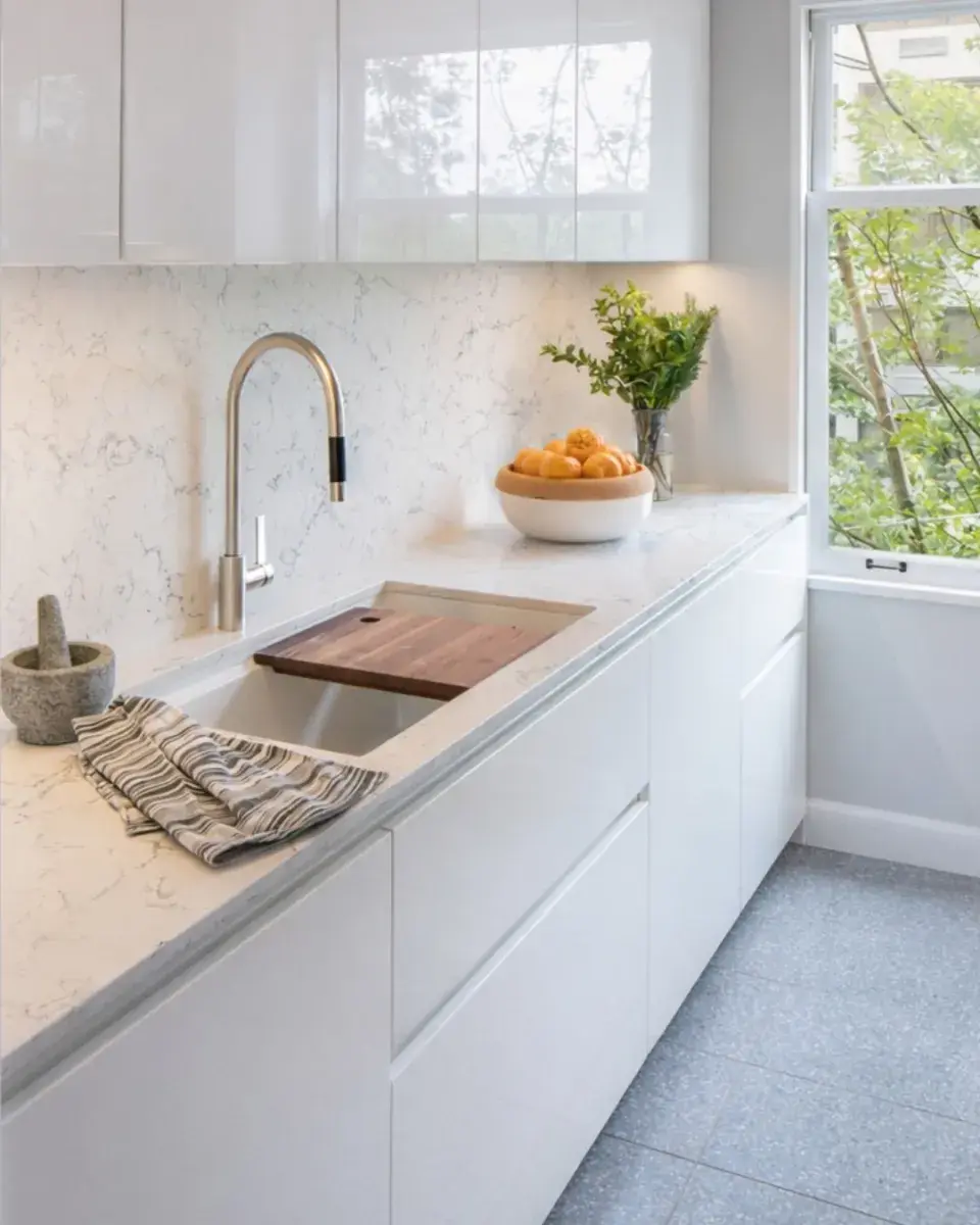 Modern white kitchen countertop with stainless steel faucet, wooden cutting board, bowl of oranges, and window view.