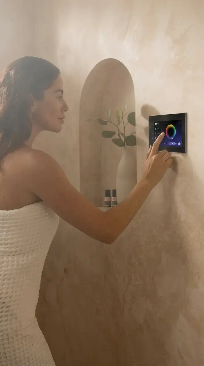 Woman in a towel adjusting color settings on a wall-mounted touchscreen in a neutral-toned bathroom.