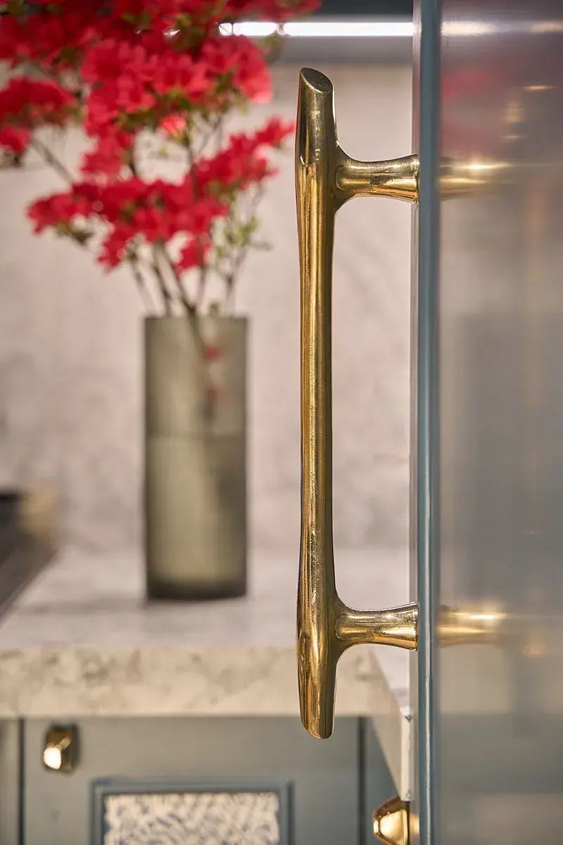 Close-up of a shiny brass door handle on a muted blue door with red flowers blurred in the background.