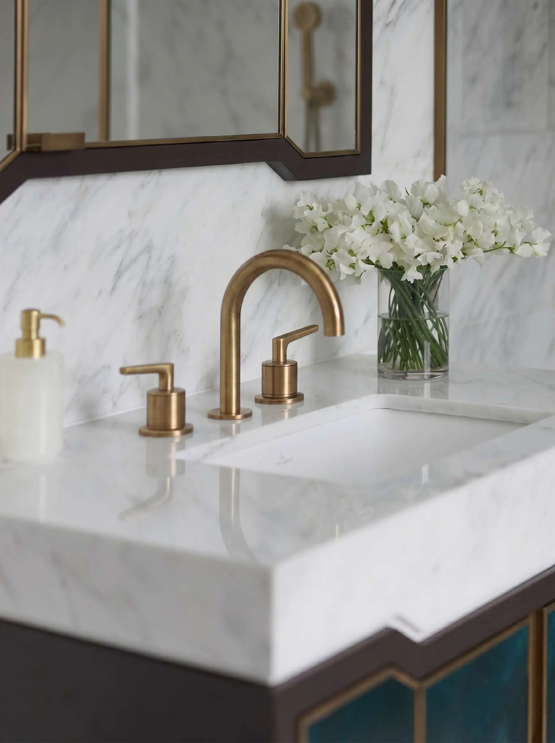 Brushed brass THG faucet and hardware on a white marble bathroom vanity with a glass vase of white sweet pea flowers.